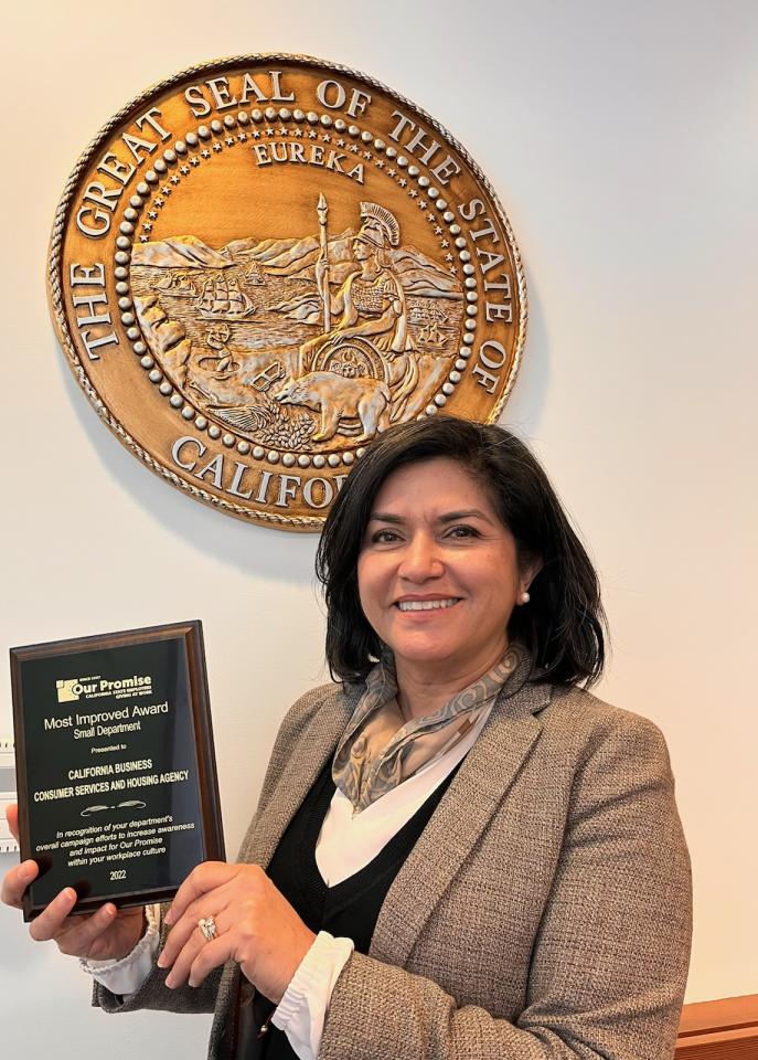 Latina woman wearing business attire holding an award  plaque with the seal of California in the background.