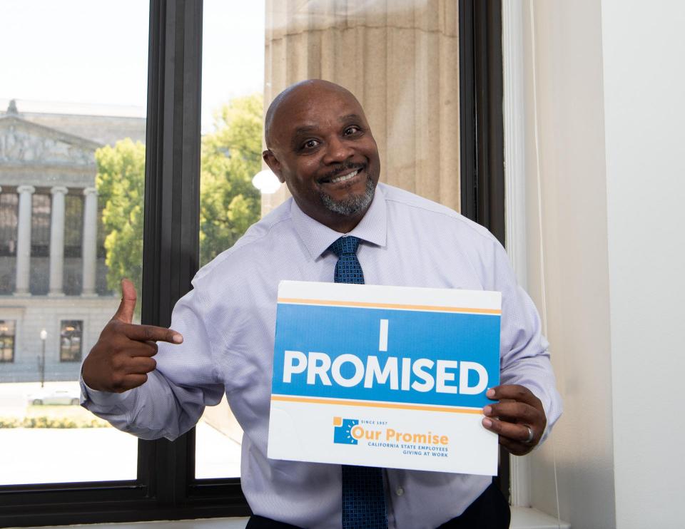Black man wearing a collared shirt and tie smiling, pointing to a sign that reads, "I PROMISED."