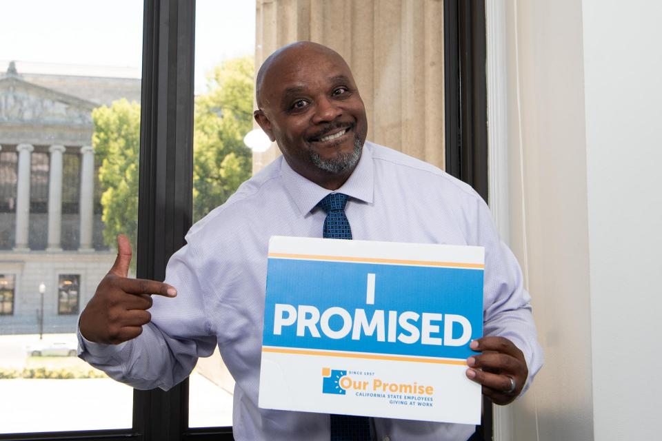 Black man wearing a collared shirt and tie smiling, pointing to a sign that reads, "I PROMISED."
